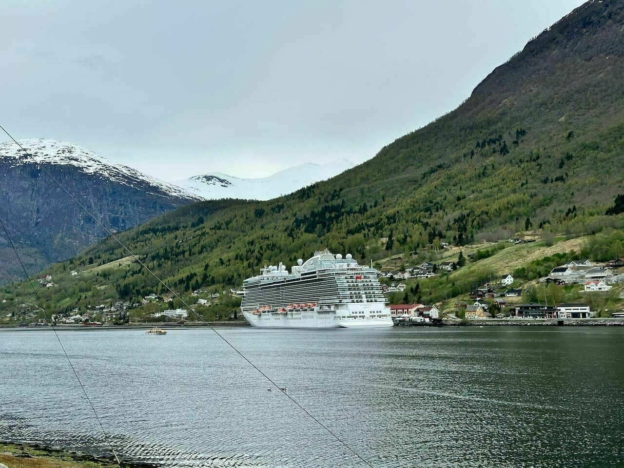 Auto-generated description: A large cruise ship is docked at a scenic fjord surrounded by lush green mountains with a few snow-capped peaks in the background.