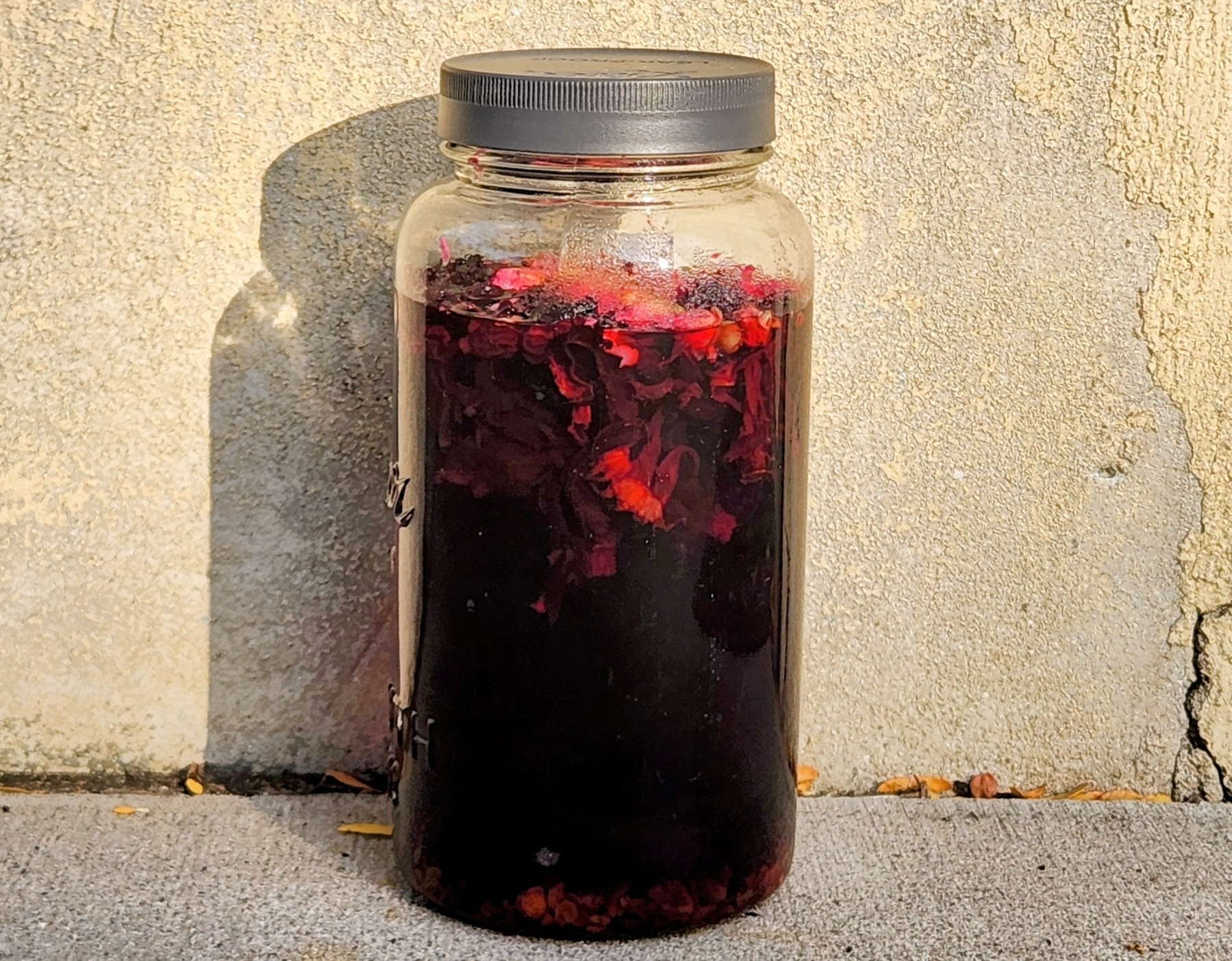 Tall mason jar of ruby red sun tea brewing against a tan stucco wall 