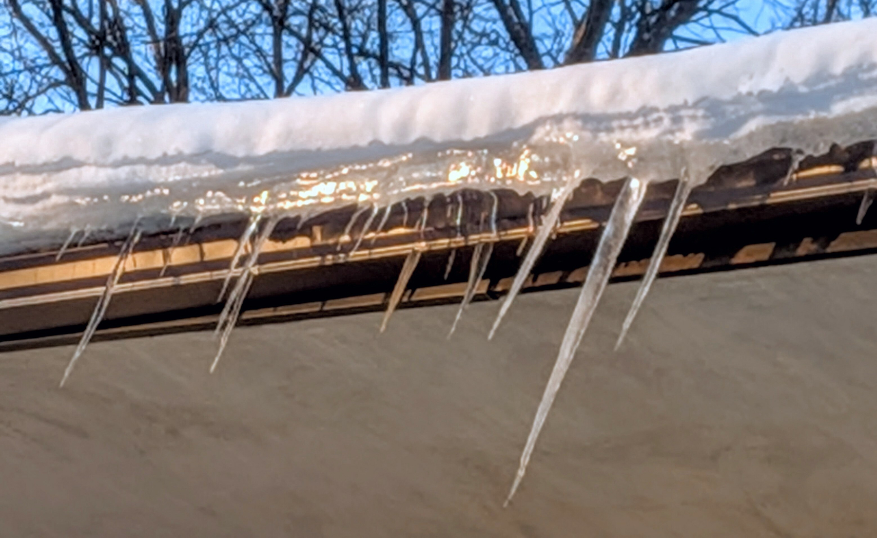 Roofline of snow, ice, and icicles beneath blue sky and dark tree limbs.