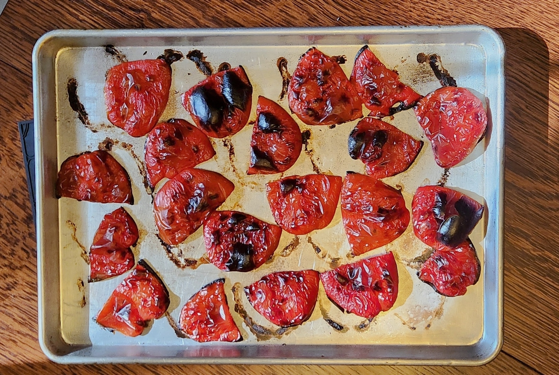 Sheet pan laid out with roasted peppers, deep red with black blisters, against a wooden table 