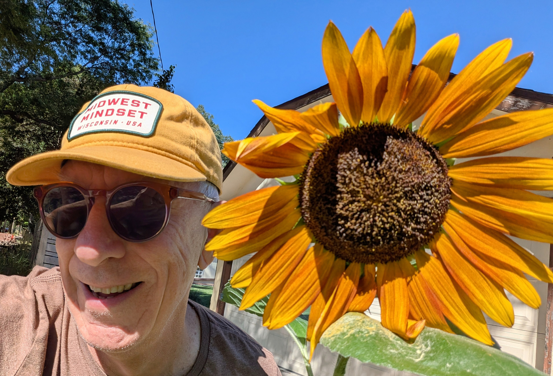 Smiling face of man with yellow ball cap and sunglasses next to a large orange sunflower bloom.
