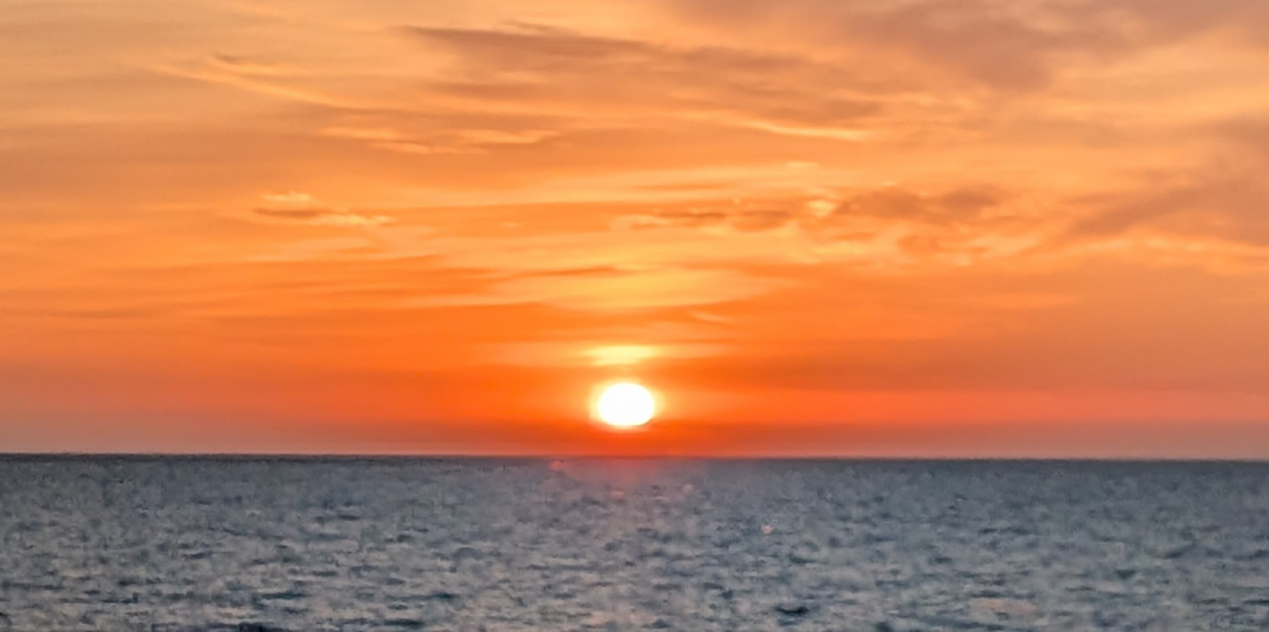 Sunrise over a lake of dark blue water with brilliant layers of red, orange, and yellow intermixed by wispy gray clouds. A postcard picture with the white oval sun just hanging above the water's horizon.