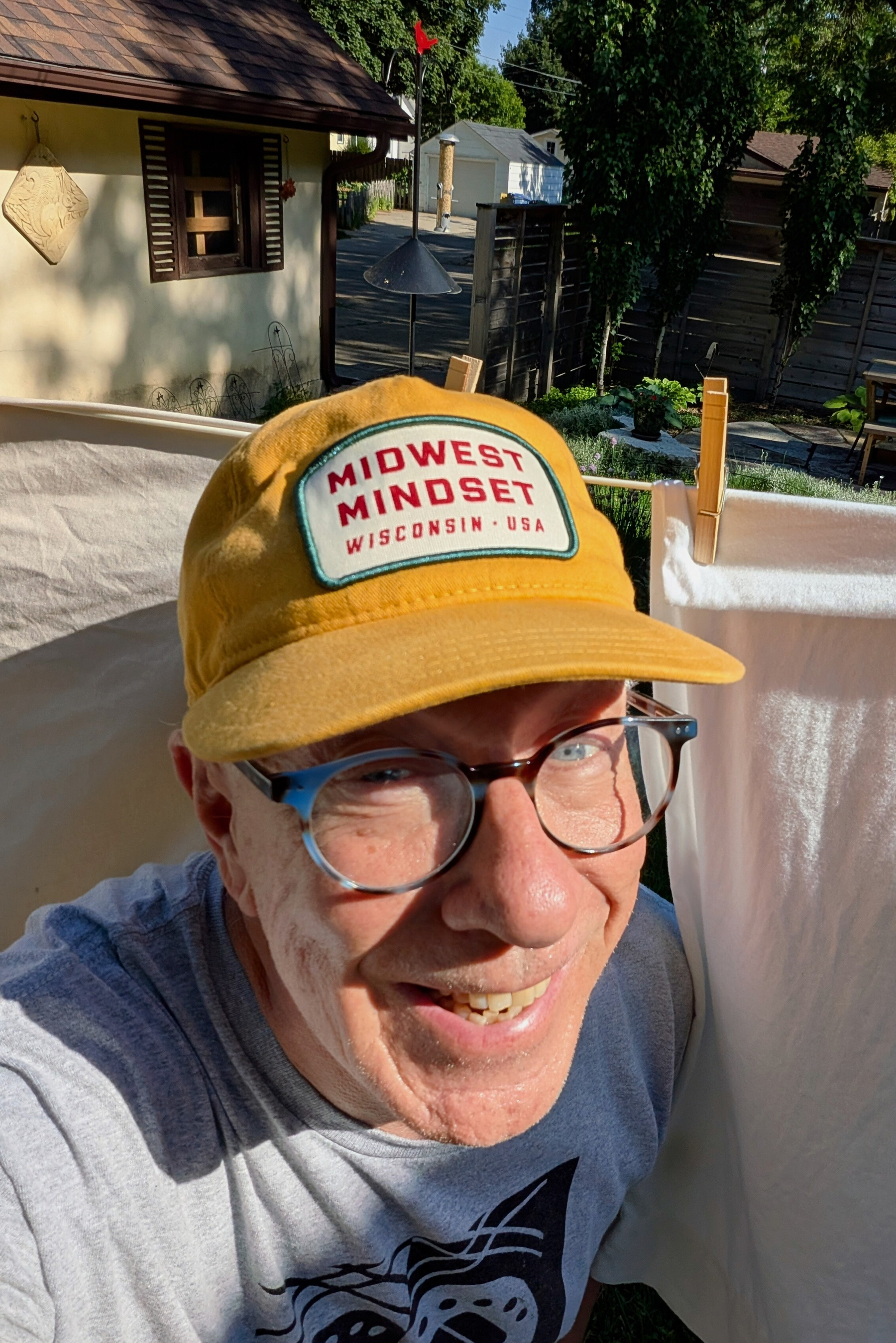 White man with yellow ball cap reading MIDWEST MINDSET against a clothesline hanging sheets outside with sunny yard background.