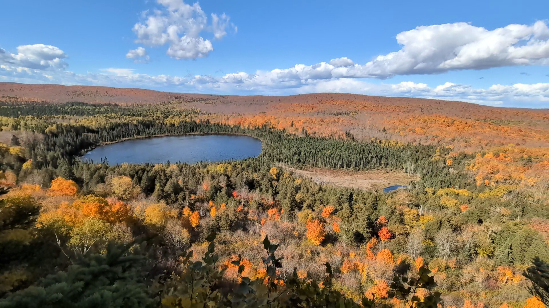 Postcard landscape of a long scenic view of an autumn forest with green, yellow, orange, and red colors rolling across hills, and a small lake reflecting a blue sky with some small puffy clouds on a sunny day.