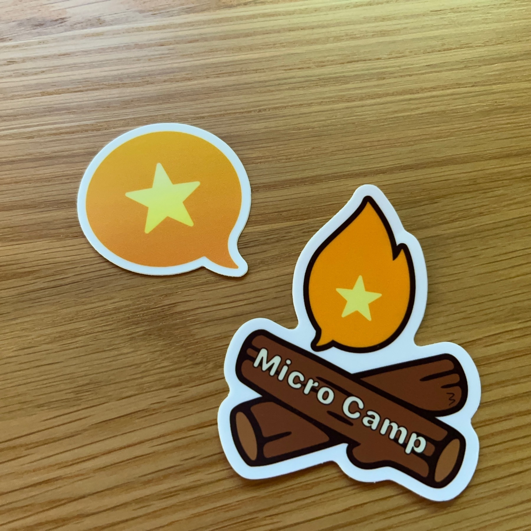 micro.blog sticker and Micro Camp logo of two logs and a fire on a sticker on a wooden tabletop. 