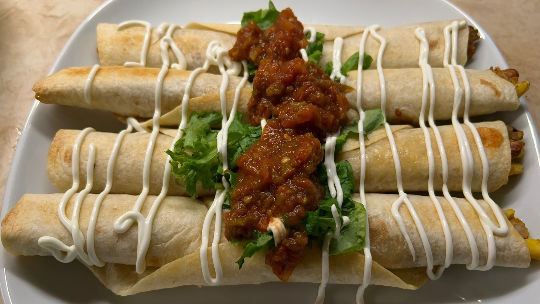 Four rolled tortillas lined up side by side with lines of sour cream, shredded lettuce, and line of red salsa down the middle.