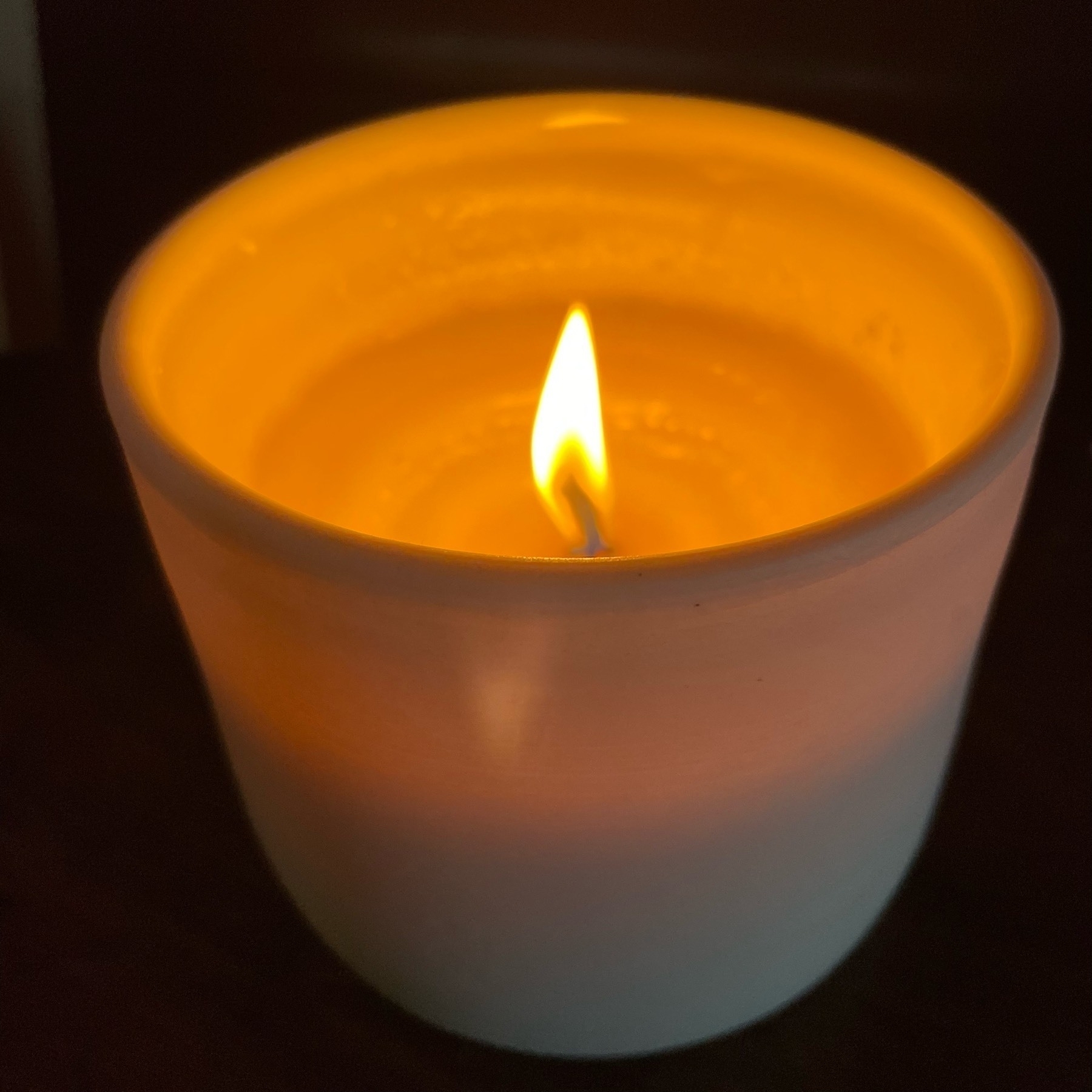 A candle flame burning on a white candle in a white circular porcelain candle holder. 