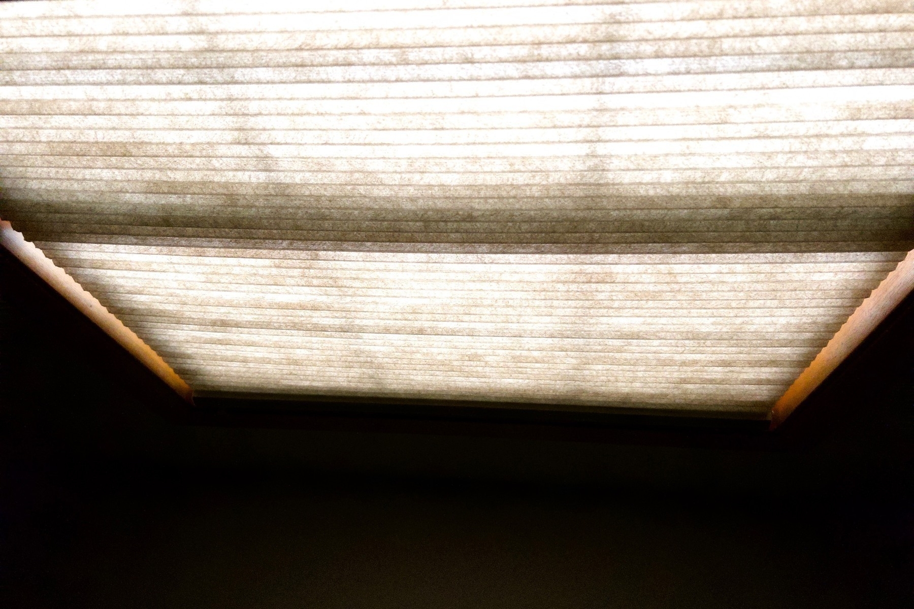 Light through a beige cellular cloth blind over a window viewed from below. 