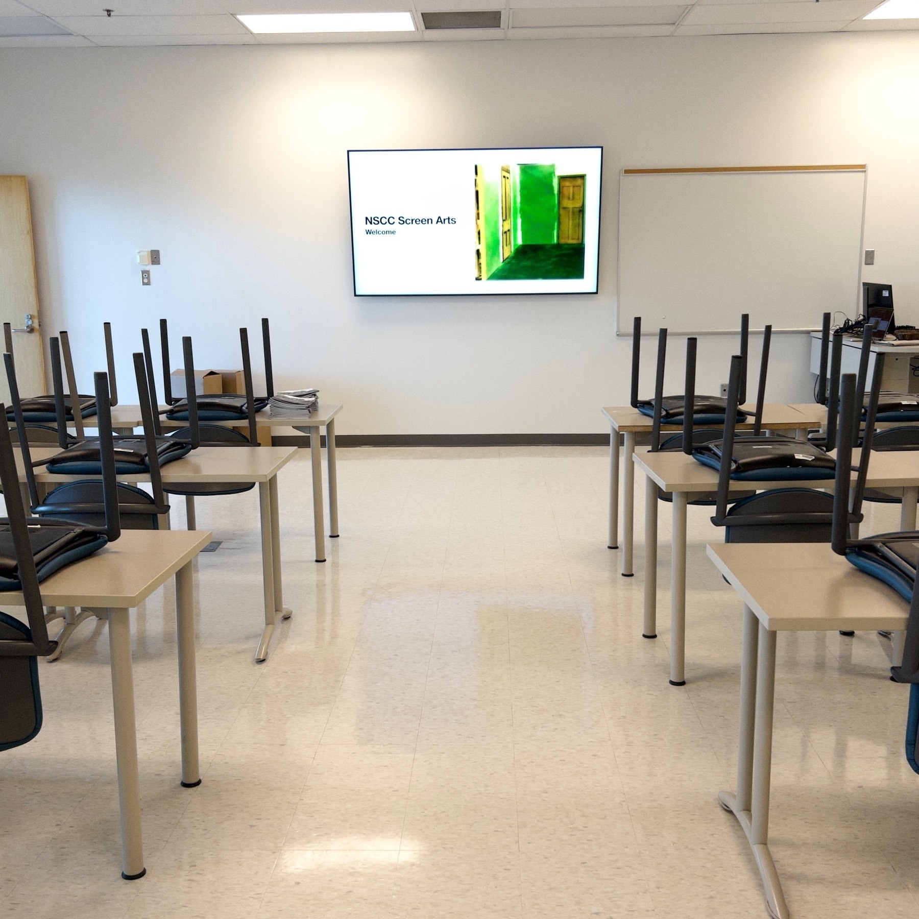 Classroom with tables and chairs upside down on top of them with a television screen with "Welcome" and "Screen Arts" written on it. 