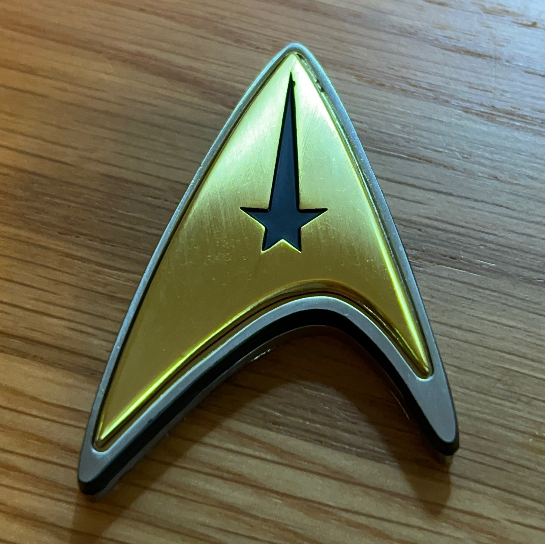 Star Trek: Strange New Worlds gold delta shaped metal command badge on a wooden table. 