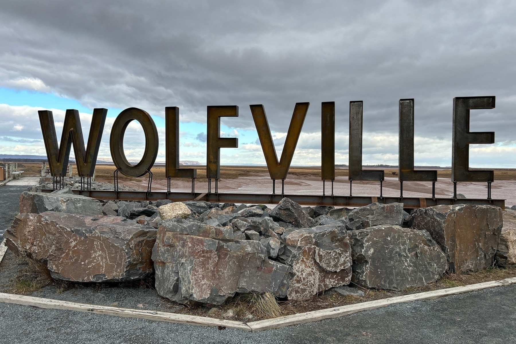 Metal artwork of the letters spelling Wolfville with a sidewalk in the foreground and clouds in the sky behind it. 