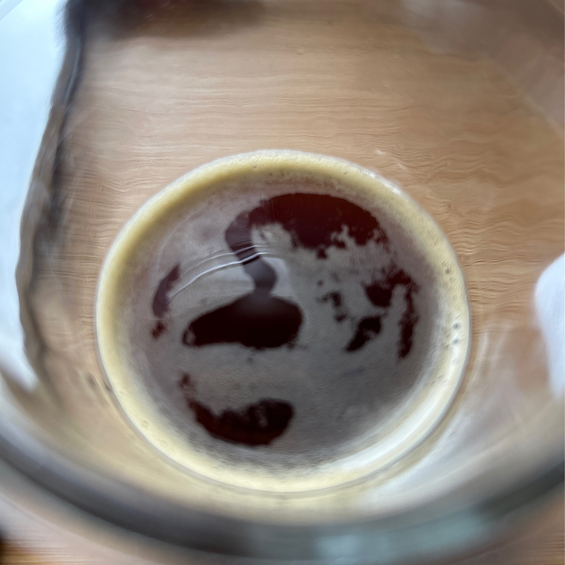 Looking down at espresso in the bottom of a clear glass on a wooden desk. 