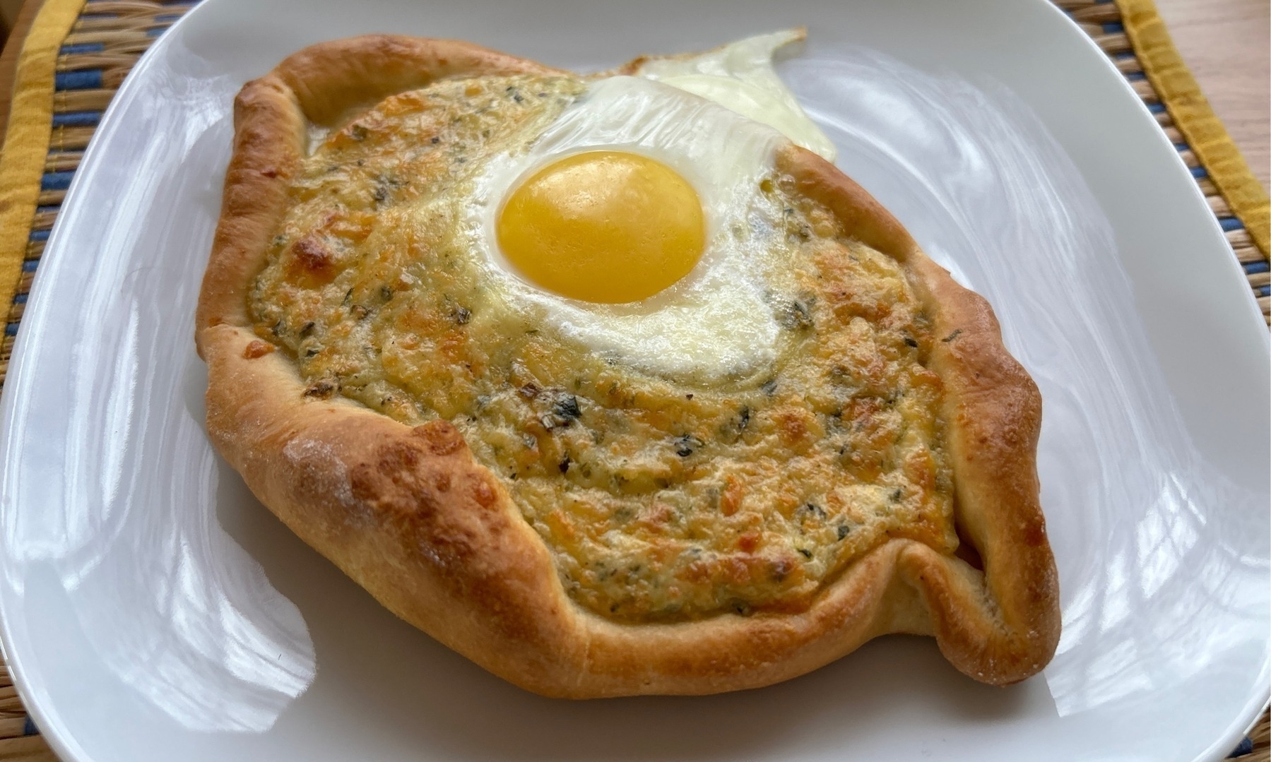 Oval bread base with a cheese filling and egg on top with everything resting on a square white plate. 