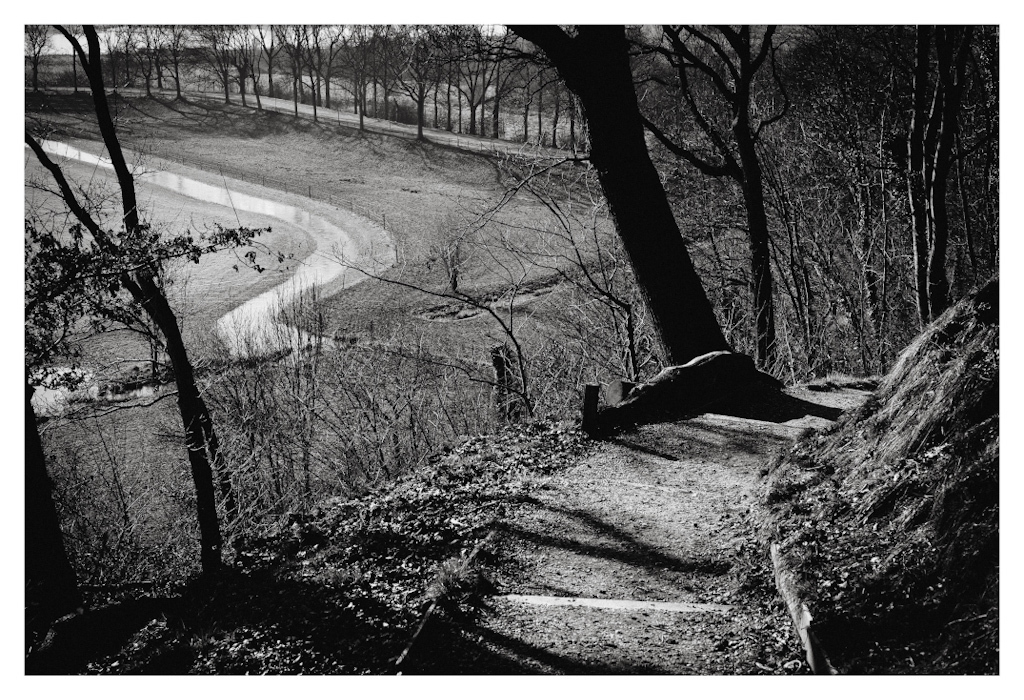 A path down a hill bending right, and a stream in the distance bending left. In black and white. 