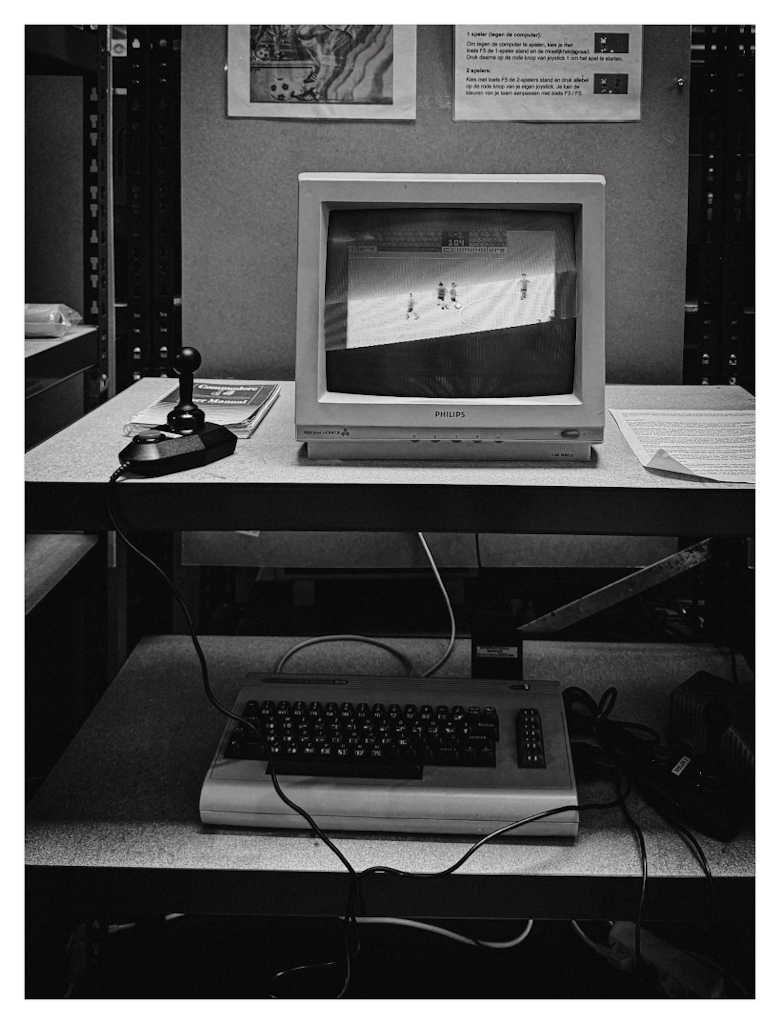 A Conmodore 64 computer on a shelf in the Bonami Games & Computers museum, with two controllers attached. Showing the game International Soccer on the connected screen, ready to play. In black and white. 