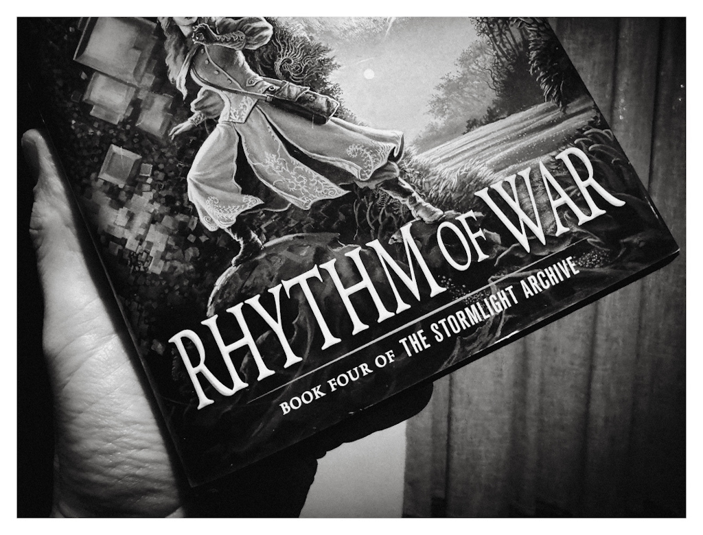 A hand holding the hardcover edition of Rhythm of War by Brandon Sanderson, in black and white. Cover art by Michael Whelan. 