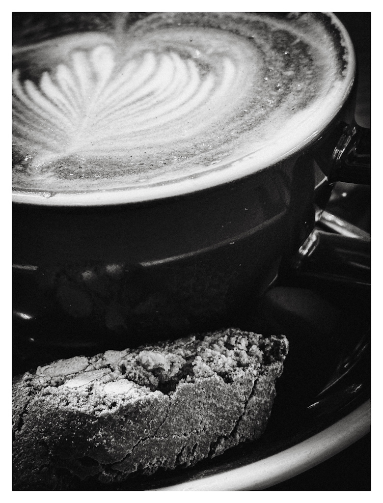 A cantucci Italian biscuit in the foreground, with a partially visible cup filled with milk foam and coffee. In black and white. 