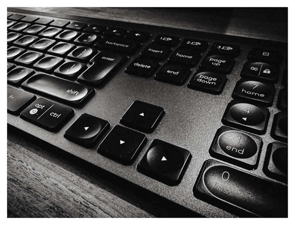 Detail of a keyboard, with the up, down, left and right arrows in the centre. The key caps are highlighted by light coming in through a window. In black and white. 