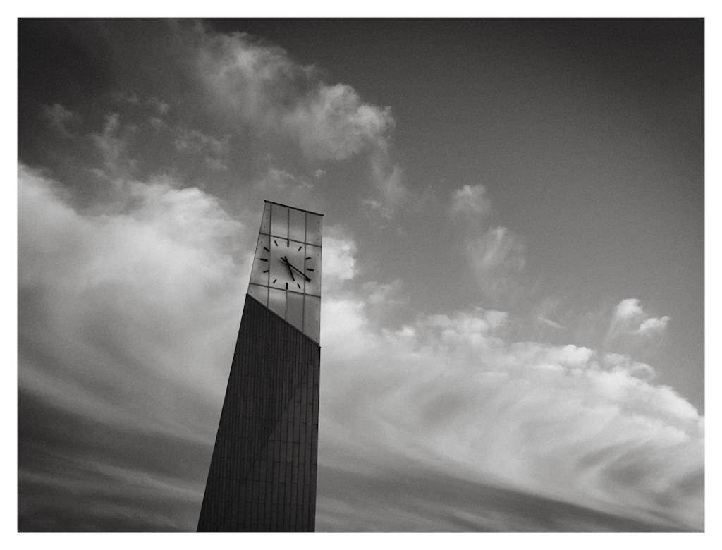 A cloud in the shape of a rolling wave brushing past a modern tower with a minimalist clock on the facade. In black and white. 