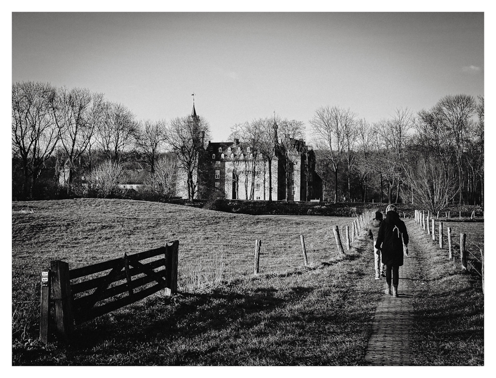 A wooden fence in the foreground, and people walking on a path towards a castle in the distance. In black and white. 