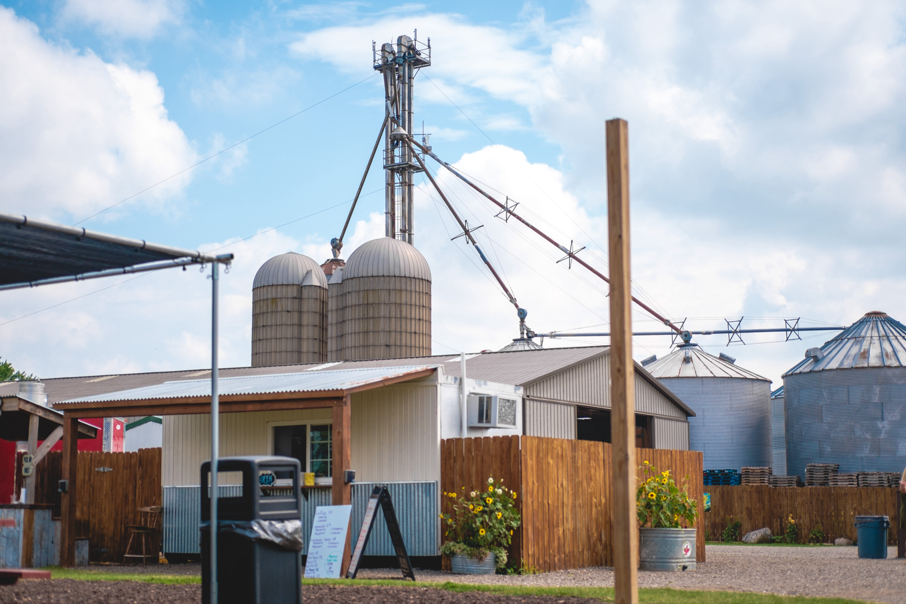 A farm with silos, a grain elevator, and various agricultural buildings under a blue sky with clouds. There’s a wooden fence and sunflowers in the foreground.