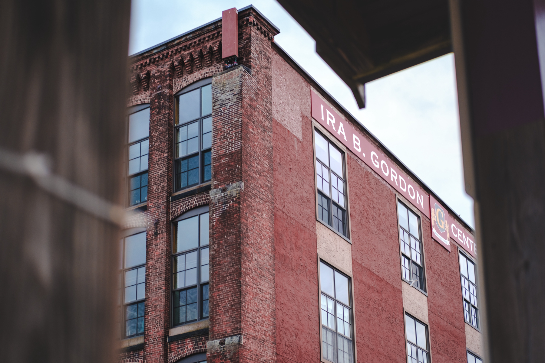 An upward shot of an industrial brick building with large windows and a sign that reads “IRA B. GORDON CENTER” framed through a wooden structure 