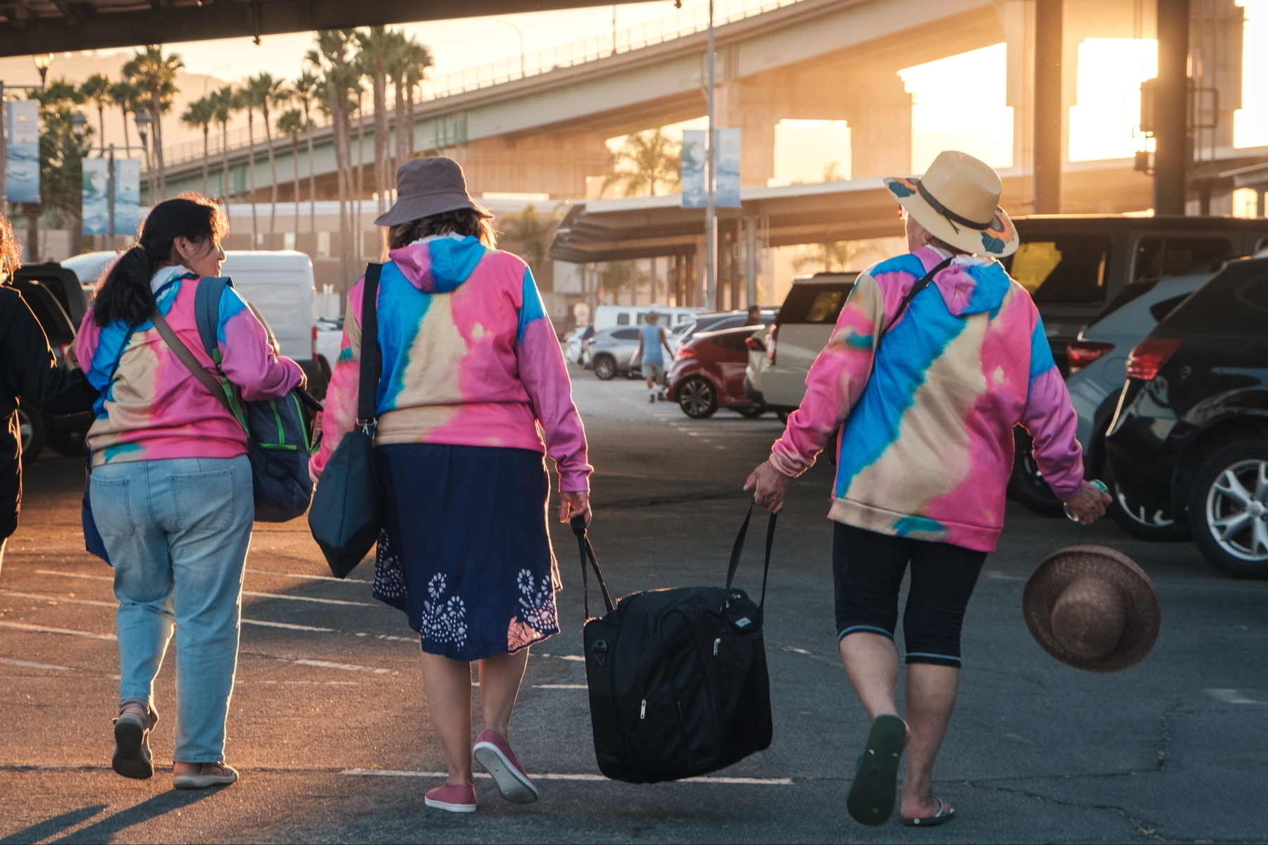 Three people wearing colorful tie-dye hoodies and hats are walking through a parking lot. They are carrying bags and one person has a hat in hand. The sun is setting, casting a warm glow over the scene with palm trees and a bridge in the background. 