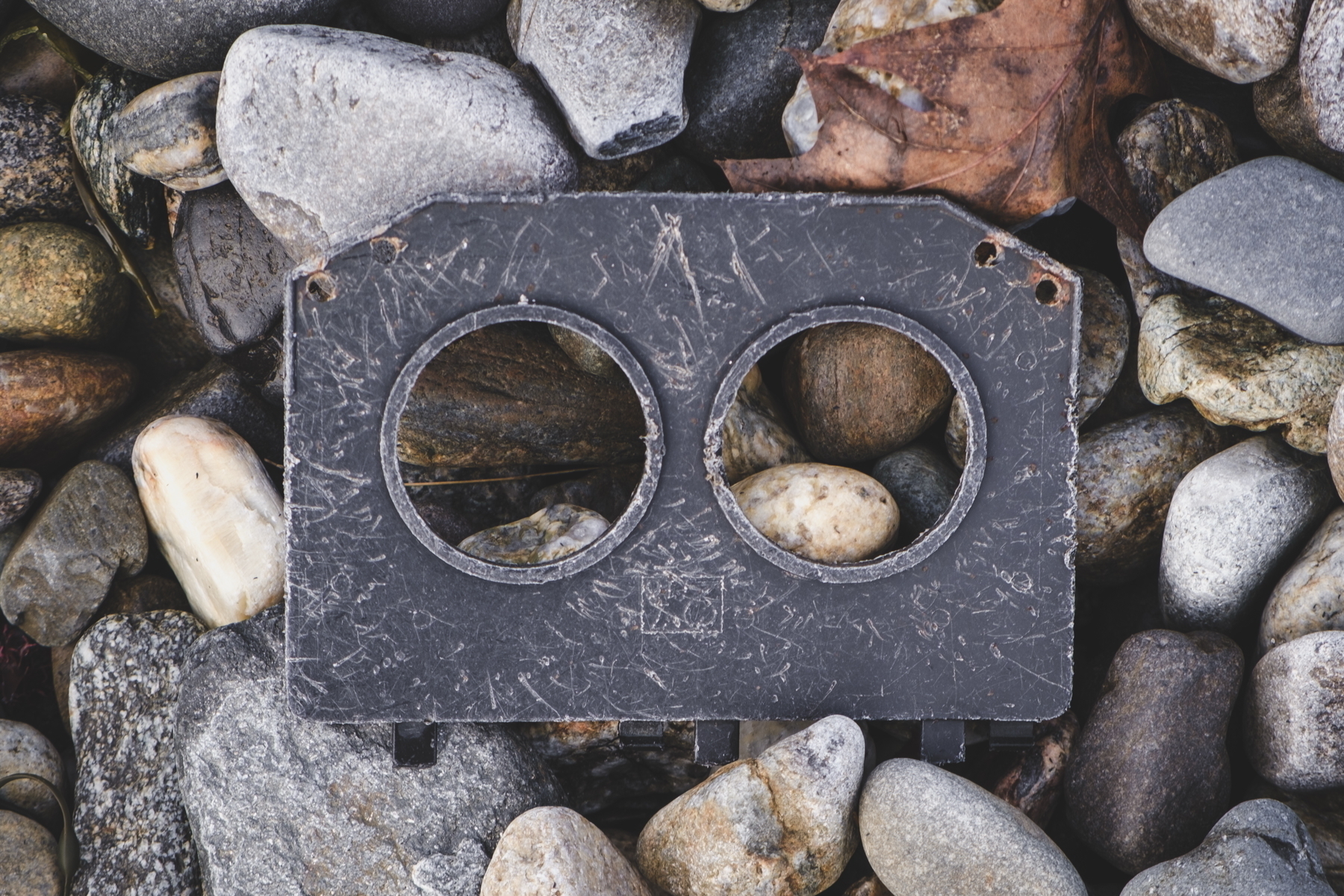 A weathered, black rectangular metal piece with two circular holes lies on a bed of smooth stones.