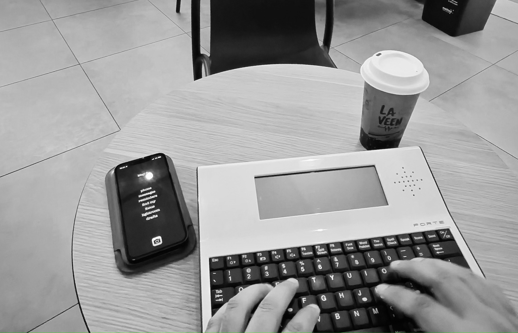 Black and white photo of an iPhone 13 Mini on a table next to a Writer Forte electronic word processor and a takeaway coffee