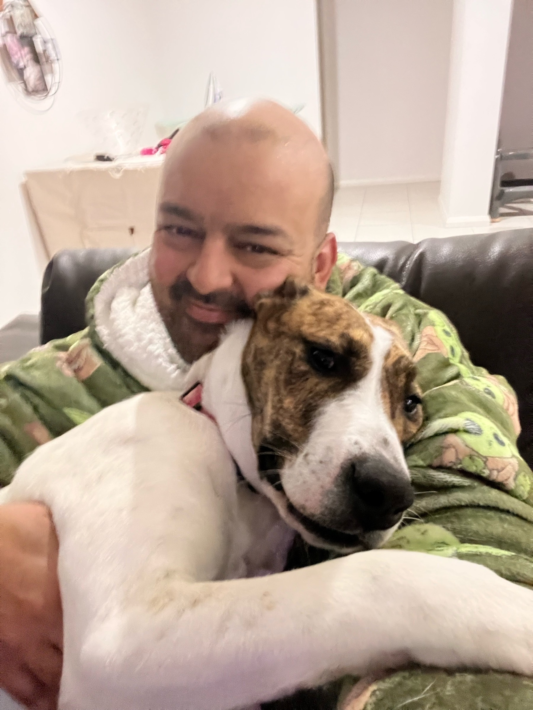 A man cuddling with a large white and brown dog indoors, wearing a green robe with patterns.