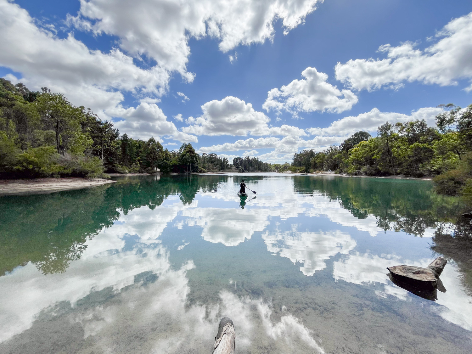 A person paddleboarding on a calm lake with clear reflections of clouds and surrounding forest - image by author