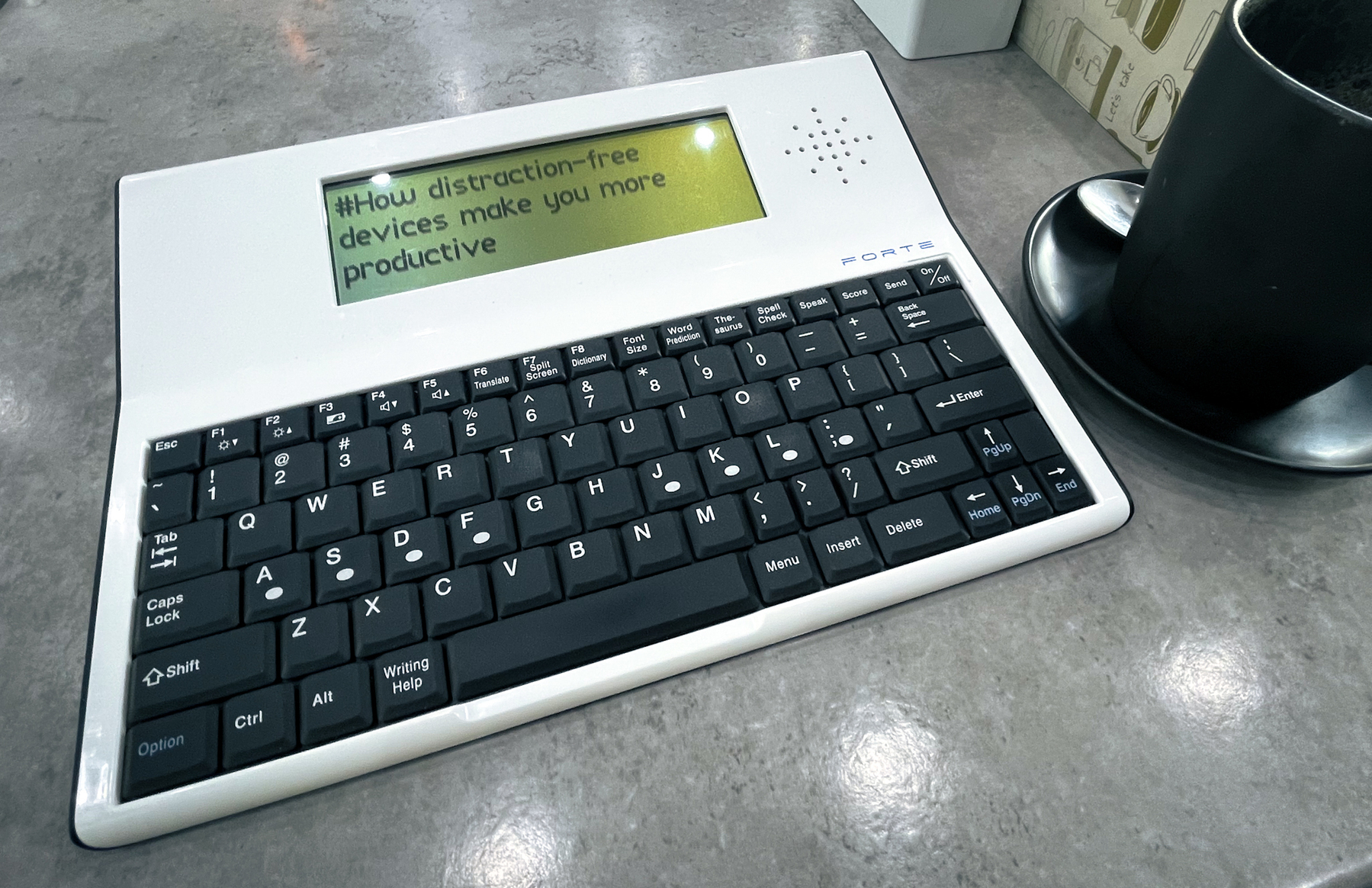 Auto-generated description: A portable distraction-free writing device with an electronic screen is displayed on a table next to a black coffee cup.