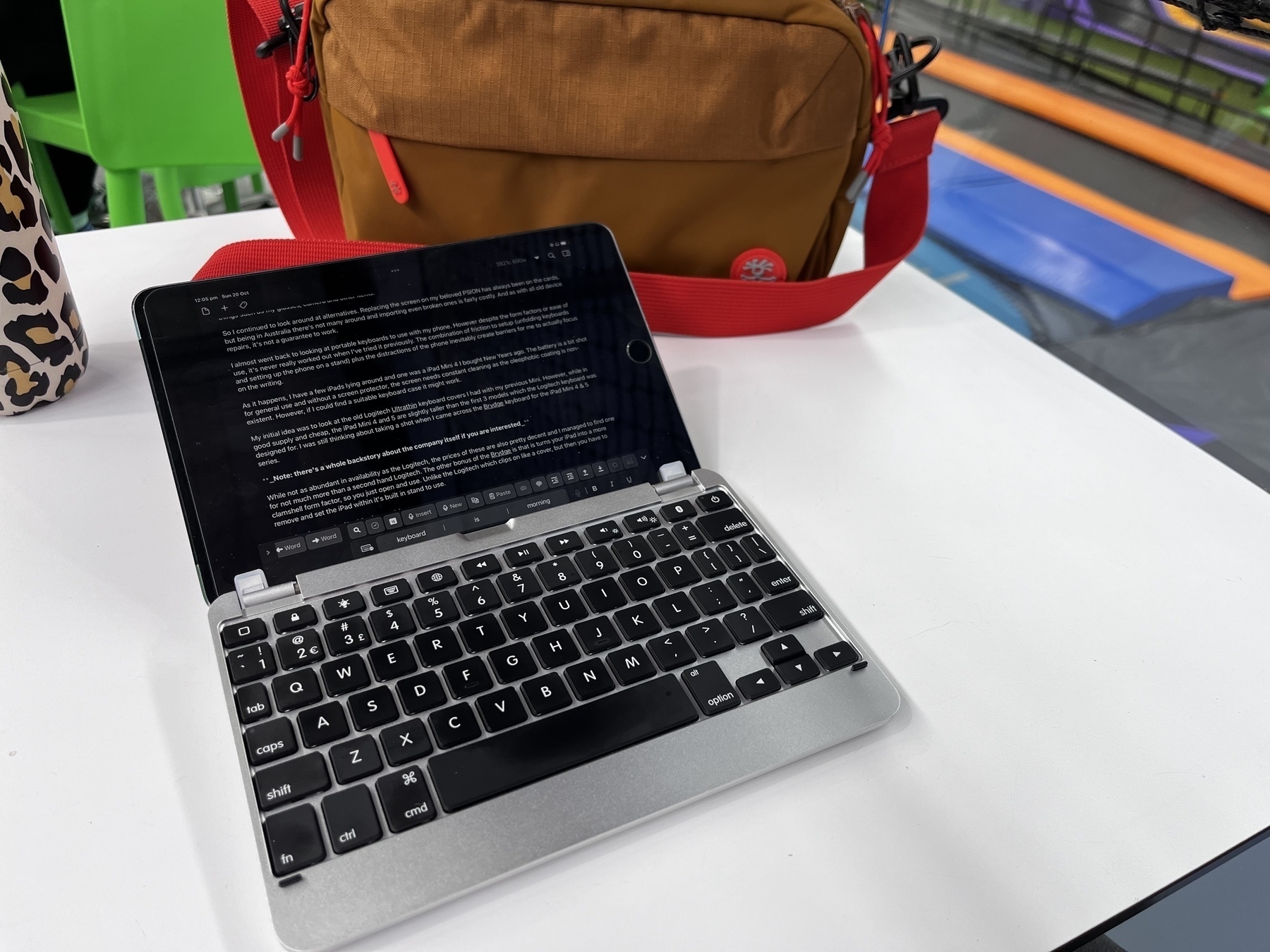 Auto-generated description: A tablet with a keyboard case sits on a white table next to a brown bag with a red strap.