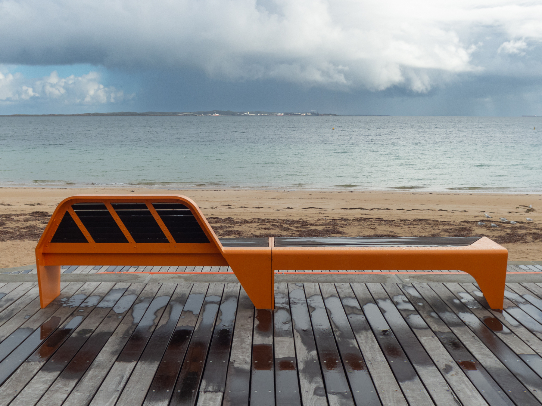 Auto-generated description: An orange bench is positioned on a wooden boardwalk overlooking a sandy beach and a cloudy seascape.