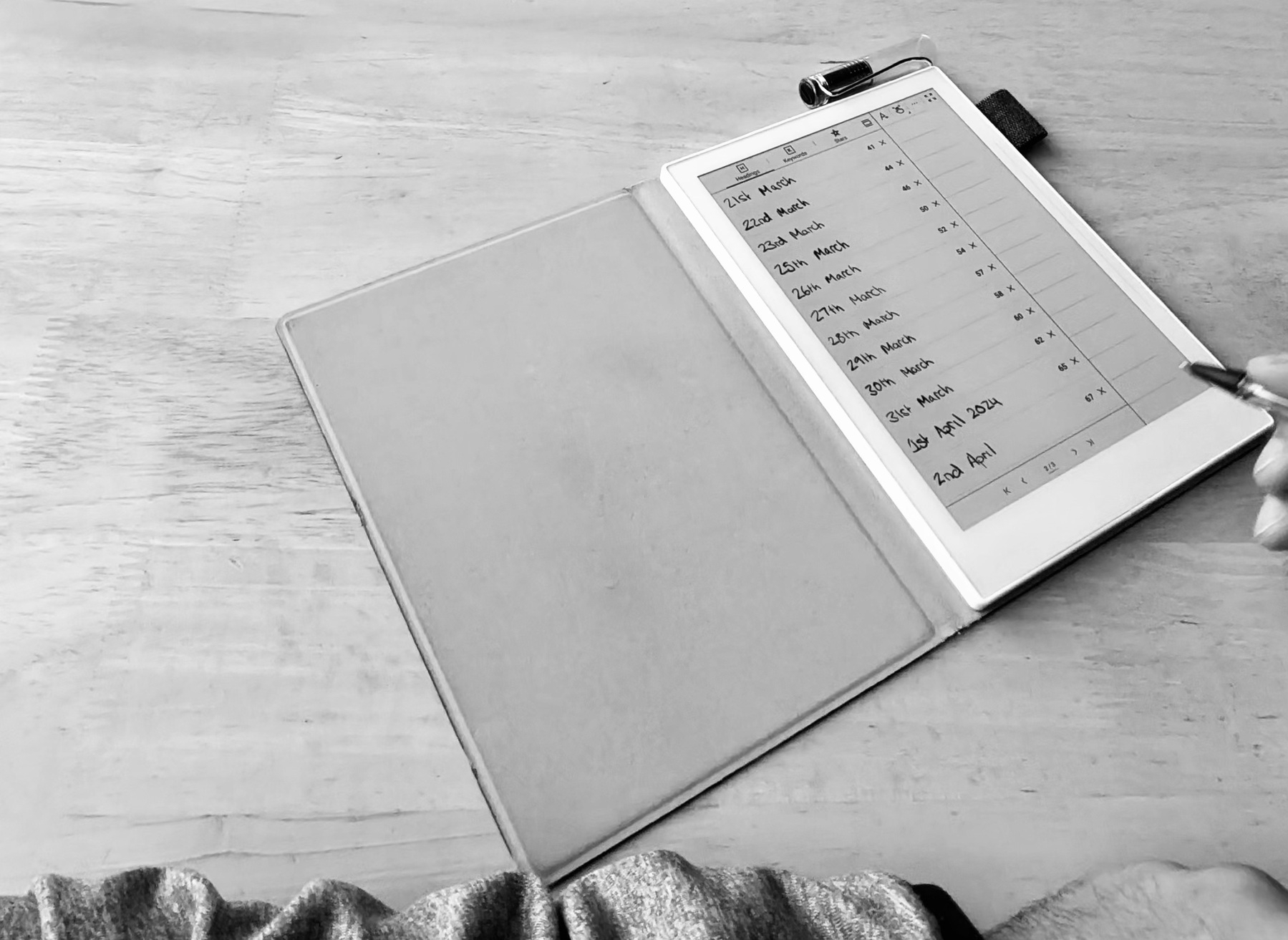 An open notebook with a handwritten list, next to a tablet displaying a calendar, on a wooden surface.