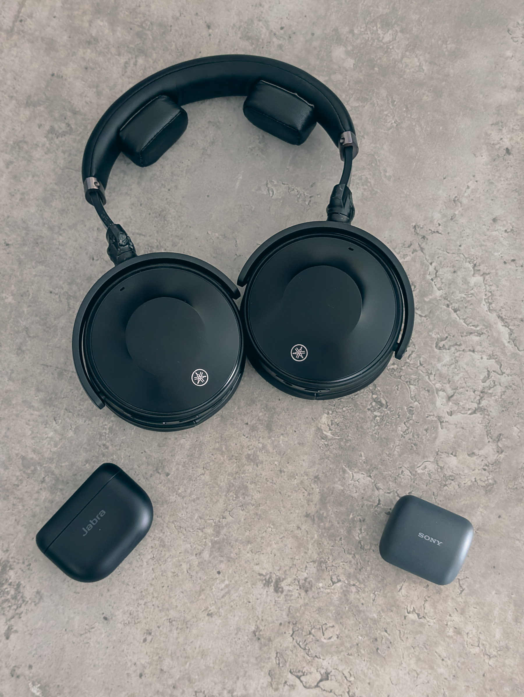 Auto-generated description: A pair of black over-ear headphones is placed on a gray surface alongside two wireless earbud cases from Jabra and Sony.