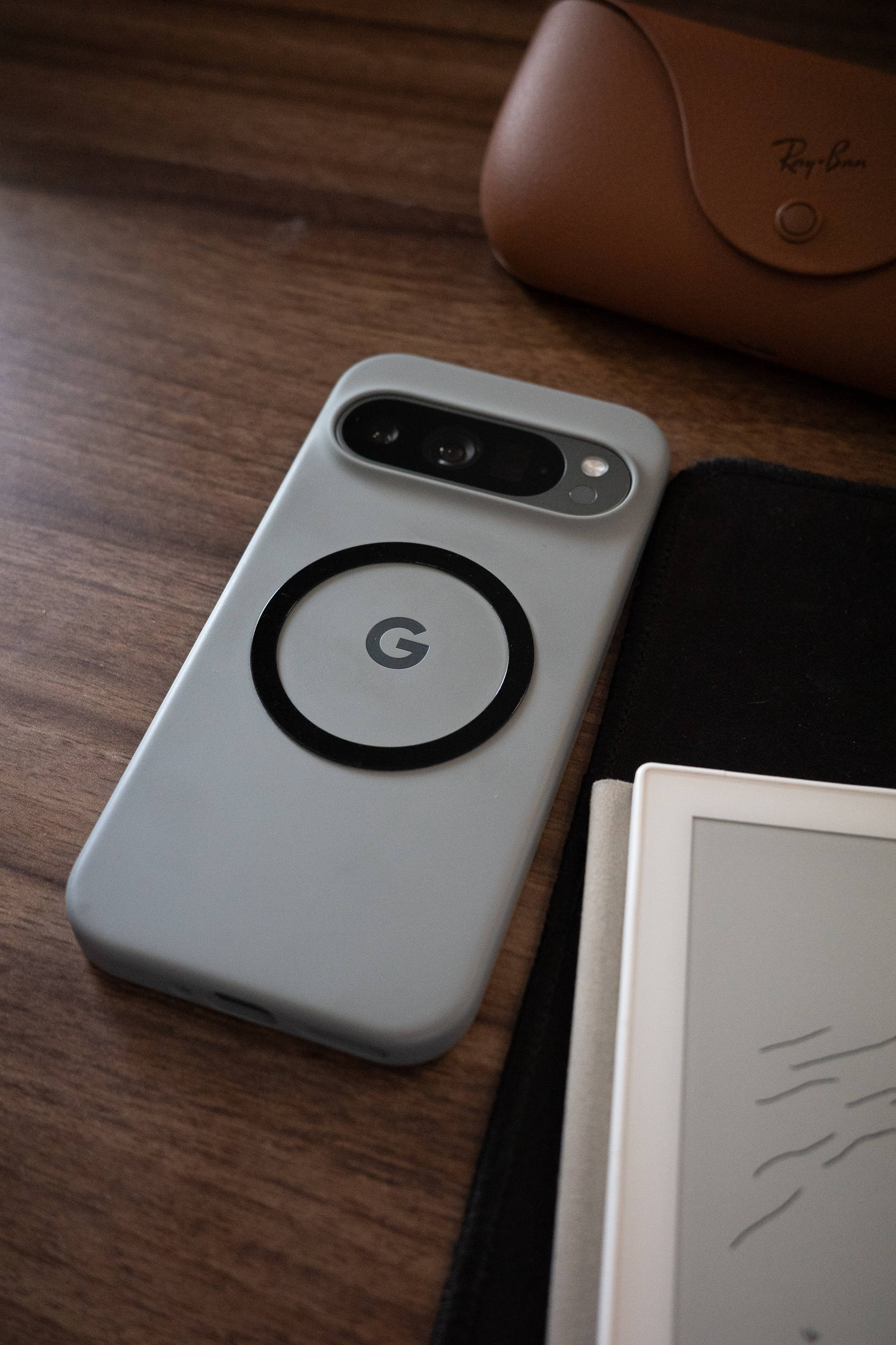 Auto-generated description: A smartphone with a gray case featuring a G logo is placed on a wooden surface next to a pair of glasses in a brown case and an electronic device with a screen.