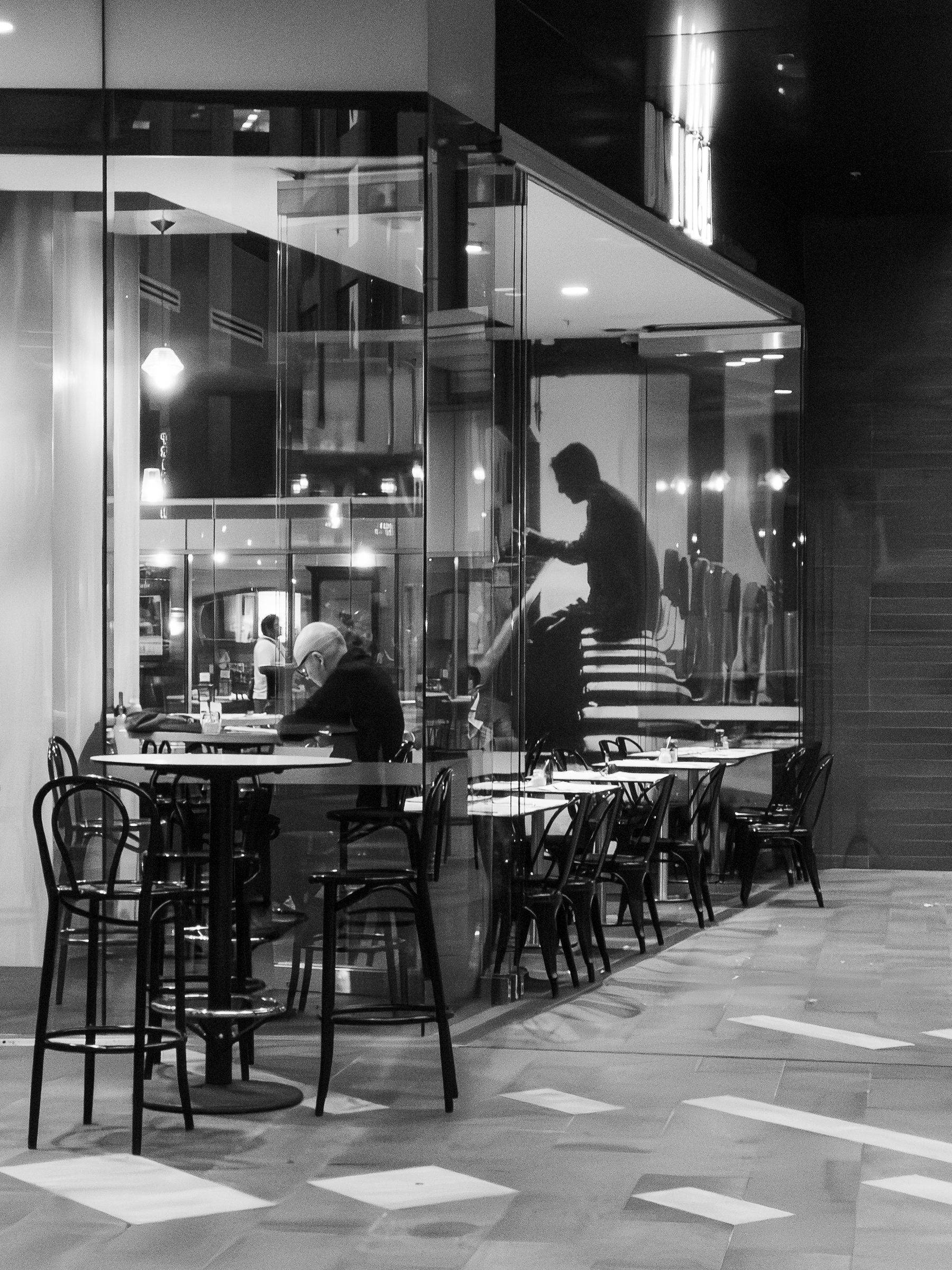 Auto-generated description: A person sits alone at a table in a modern cafe with empty chairs and a large window reflecting a silhouette.