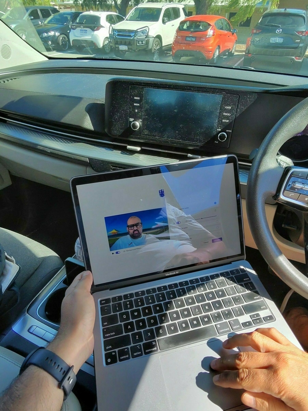 A person is sitting in a car using a laptop, with a video call on the screen and a parking lot visible through the windshield.