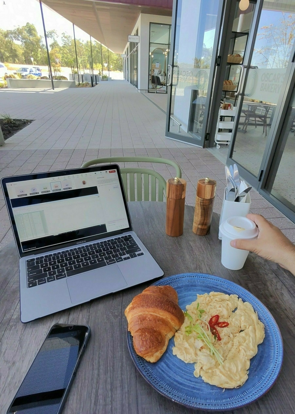 A person is enjoying a meal of scrambled eggs and croissants with coffee at an outdoor table, alongside a laptop and a phone.