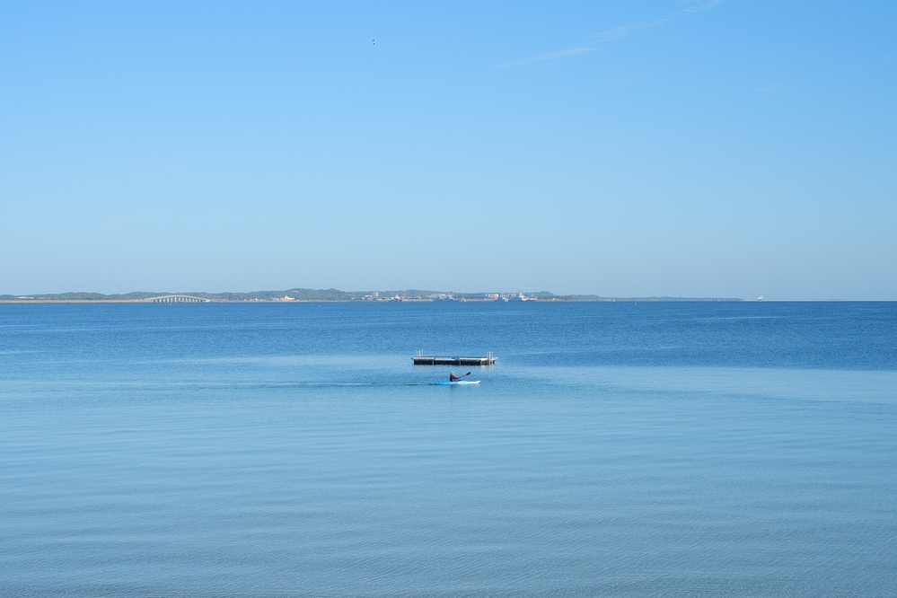 A lone kayak floats near a small platform on a calm, expansive body of water with a distant shoreline under a clear blue sky.