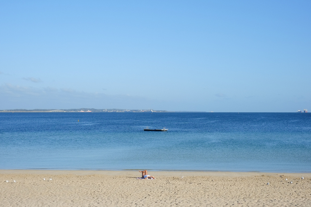 A tranquil beach scene features a sandy shoreline, a lone person lounging under an umbrella, and a calm sea stretching to the horizon with a small floating platform.