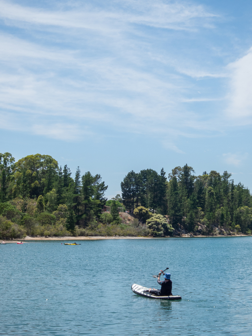 A person is kayaking on a tranquil body of water with trees lining the distant shore.