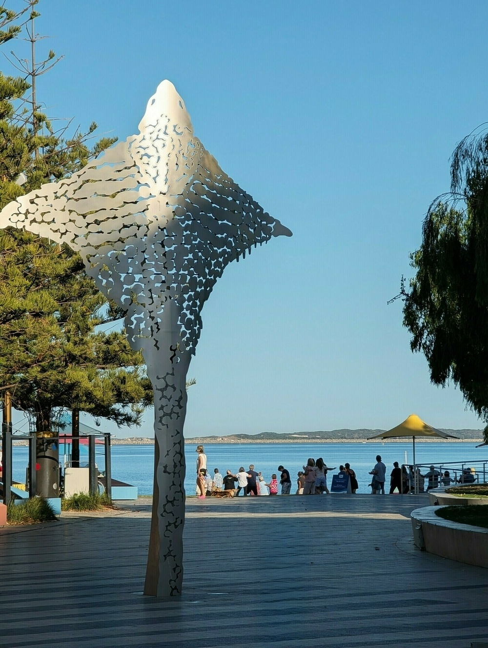 A metal sculpture resembling a manta ray is set along a waterfront with people and trees nearby.