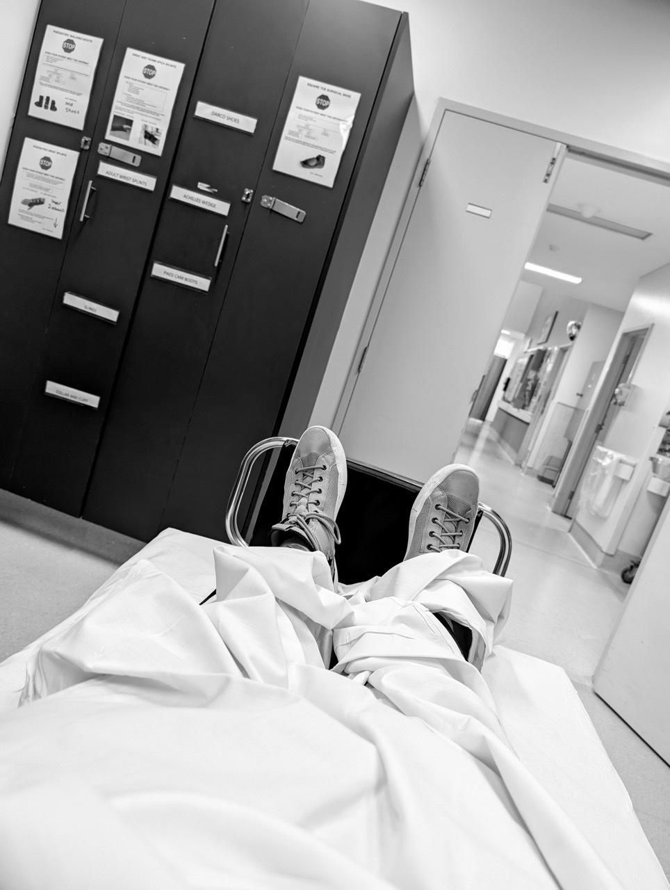 A person is lying on a stretcher in a hospital, looking towards the hallway and some lockers.