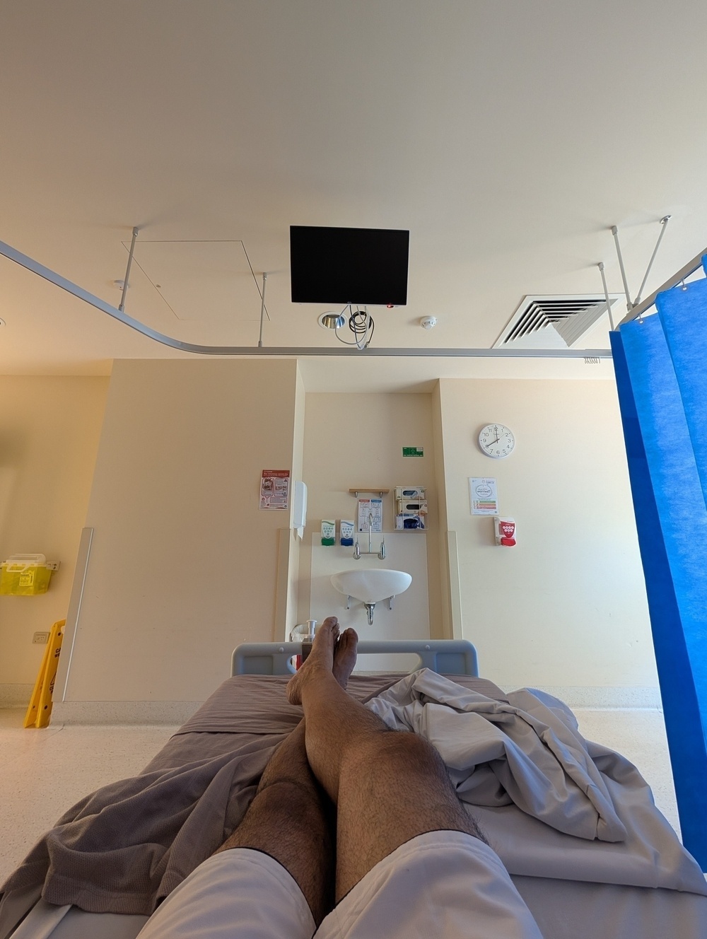 A person is lying on a hospital bed with a view of a wall-mounted sink, medical equipment, and a clock.