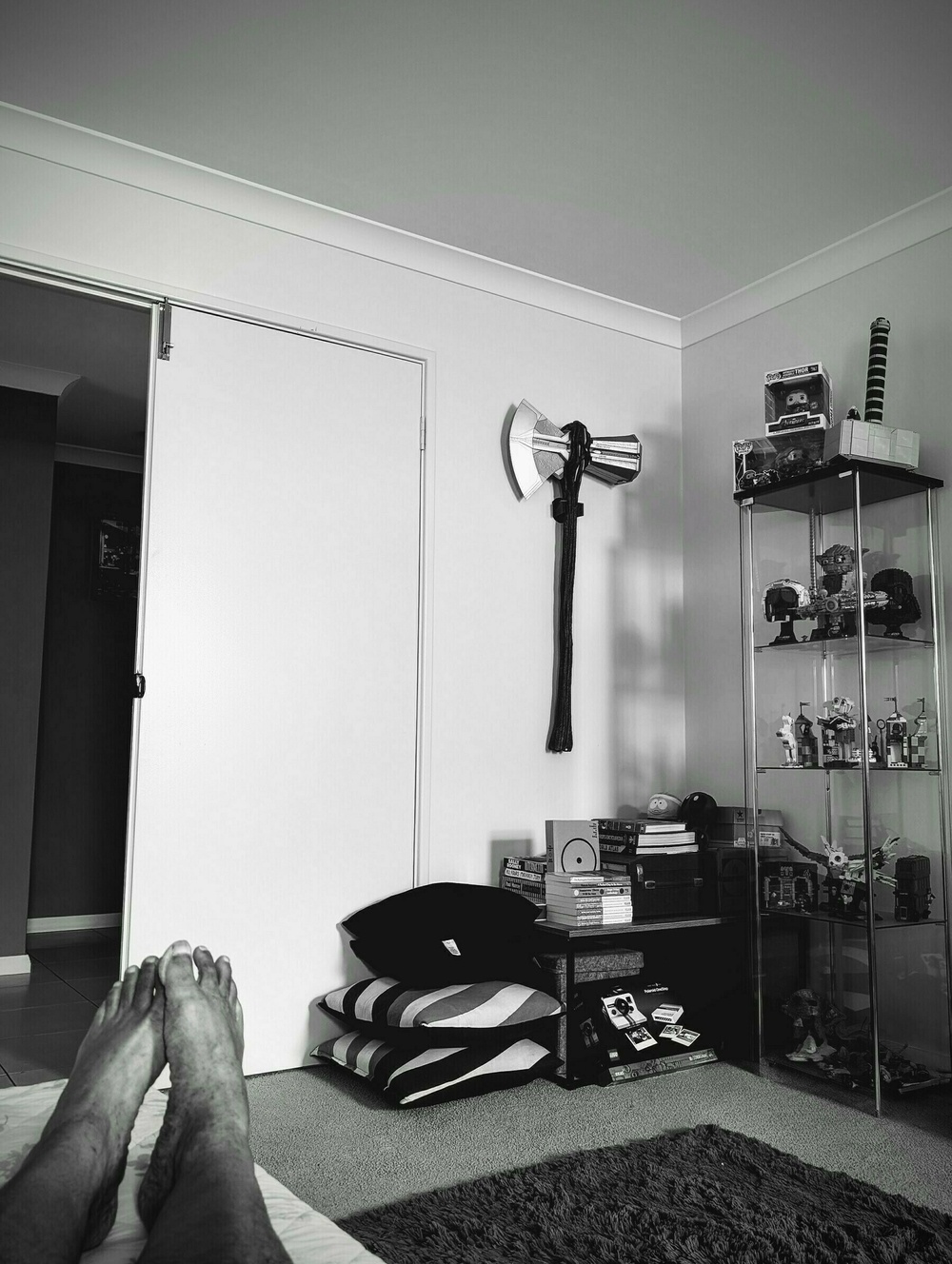 A person lying on a bed is looking at a bedroom corner with a shelf displaying collectibles, a toy axe hanging on the wall, and a small stack of pillows and books on the floor.