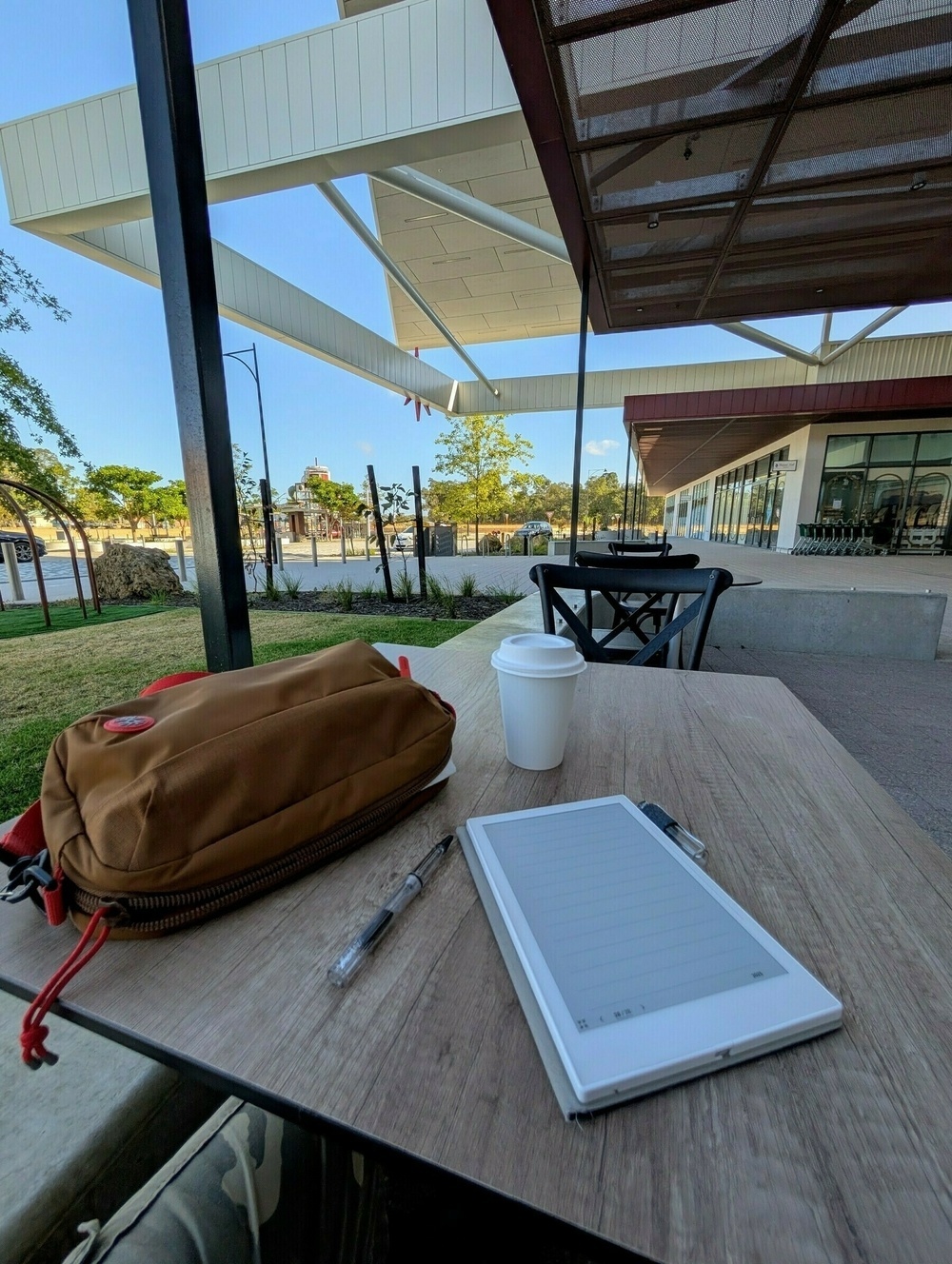 A table holds a tablet, pen, cup, and backpack, set against a modern outdoor patio with a clear sky in the background.