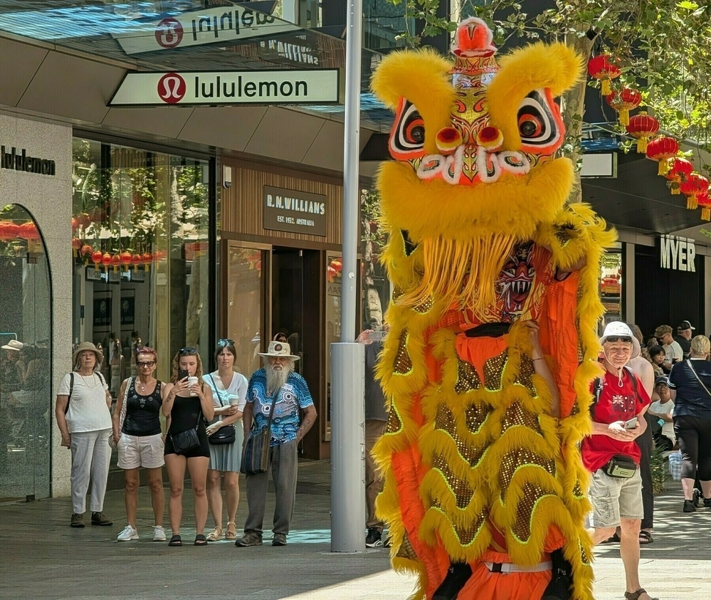 A vibrant lion dance performer in yellow and orange is entertaining a crowd outdoors near a Lululemon store.