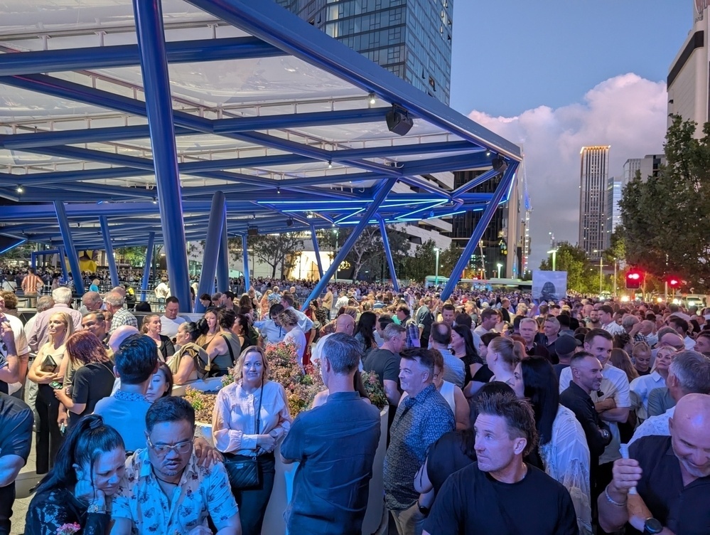 A large crowd of people is gathered at an outdoor event under a modern steel structure in an urban setting during the evening.